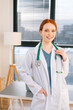 © dikushin - Portrait of friendly female doctor in white coat standing on background of window in sunny day in light medical clinic office. Young redhead woman physician posing with stethoscope looking at camera.
