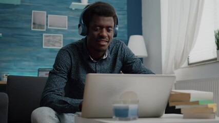 Poster - Black african american man wearing headphone, typing on laptop, using internet web online services. Remote working from home computer user using modern technology and communication devices