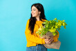 © luismolinero - Young woman holding a grocery shopping bag isolated on blue background looking side
