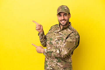 Wall Mural - Military man isolated on yellow background pointing finger to the side and presenting a product