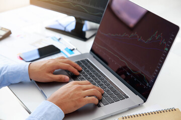  Close-up young businessman statistic graph of stock market financial indices analysis on a laptop and on a desktop computer in office. Analyzing and Trading Stock concept.