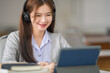 © EduLife Photos - Young Asian woman college student with wireless headphones watches and listens to the lecturer on tablet and writes on the notebook to study online by herself in classroom