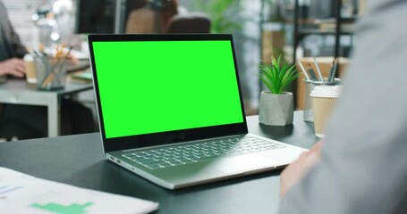Over the shoulder: creative young man sitting at his desk using laptop computer with mock-up green screen. Bright office where diverse team of young professionals working on computers