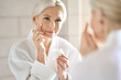 © insta_photos - Attractive mid age older adult 50 years old blonde woman wears bathrobe in bathroom applying nourishing antiage face skin care cream treatment, looking at mirror doing daily morning beauty routine.