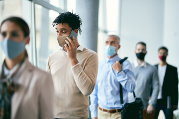 Young black businessman with face mask talking on the phone while waiting in line for job interview at office building hallway.