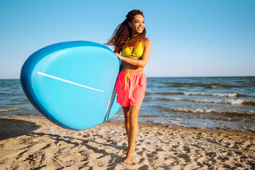 Wall Mural - Beautiful sexy woman with glanders at the beach. Woman surfer walks with a board on the sandy beach. Healthy active lifestyle. Surfing. Summer vacation. Extreme sport. Travel, weekend,  lifestyle.