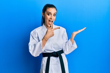 Wall Mural - Beautiful brunette young woman wearing karate fighter uniform with black belt amazed and smiling to the camera while presenting with hand and pointing with finger.