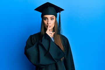 Wall Mural - Beautiful brunette young woman wearing graduation cap and ceremony robe asking to be quiet with finger on lips. silence and secret concept.