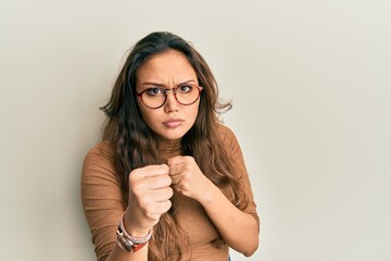 Canvas Print - Young hispanic girl wearing casual clothes and glasses ready to fight with fist defense gesture, angry and upset face, afraid of problem