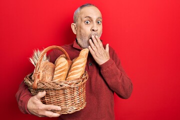 Poster - Handsome mature man holding wicker basket with bread covering mouth with hand, shocked and afraid for mistake. surprised expression