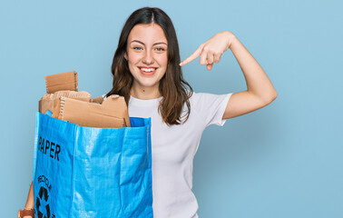 Young beautiful woman holding recycling wastebasket with paper and cardboard pointing finger to one self smiling happy and proud