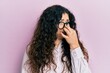 Young brunette woman with curly hair wearing casual clothes and glasses smelling something stinky and disgusting, intolerable smell, holding breath with fingers on nose. bad smell