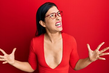 Wall Mural - Young latin woman wearing casual clothes and glasses crazy and mad shouting and yelling with aggressive expression and arms raised. frustration concept.