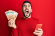 Young hispanic man using smartphone holding 50 euros banknotes angry and mad screaming frustrated and furious, shouting with anger looking up.