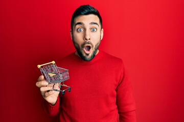 Canvas Print - Young hispanic man holding small supermarket shopping cart scared and amazed with open mouth for surprise, disbelief face