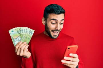 Canvas Print - Young hispanic man using smartphone holding chilean pesos banknotes smiling and laughing hard out loud because funny crazy joke.