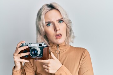 Sticker - Young blonde girl holding vintage camera in shock face, looking skeptical and sarcastic, surprised with open mouth