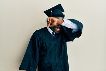 Wall Mural - Handsome black man wearing graduation cap and ceremony robe covering eyes with arm smiling cheerful and funny. blind concept.