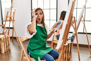 Wall Mural - Young hispanic artist women painting on canvas at art studio doing ok gesture shocked with surprised face, eye looking through fingers. unbelieving expression.