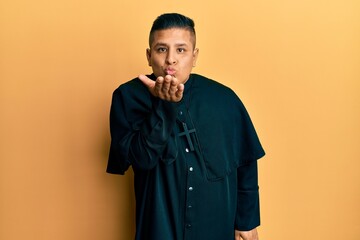 Poster - Young latin priest man standing over yellow background looking at the camera blowing a kiss with hand on air being lovely and sexy. love expression.