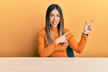 Young hispanic woman wearing casual clothes sitting on the table smiling and looking at the camera pointing with two hands and fingers to the side.