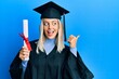 © Krakenimages.com - Beautiful blonde woman wearing graduation cap and ceremony robe holding degree pointing thumb up to the side smiling happy with open mouth