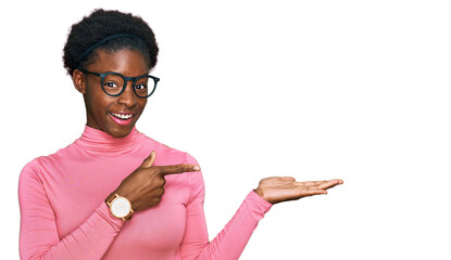 Canvas Print - Young african american girl wearing casual clothes and glasses amazed and smiling to the camera while presenting with hand and pointing with finger.