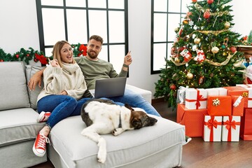 Canvas Print - Young couple using laptop sitting by christmas tree with a big smile on face, pointing with hand finger to the side looking at the camera.