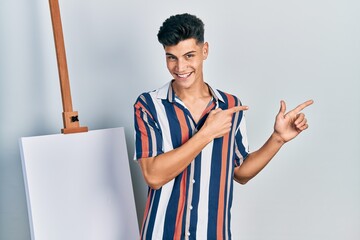 Canvas Print - Young hispanic man standing close to empty canvas smiling and looking at the camera pointing with two hands and fingers to the side.