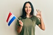 Young hispanic girl holding holland flag doing ok sign with fingers, smiling friendly gesturing excellent symbol