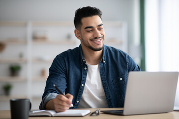 Cheerful arab guy working on laptop, taking notes