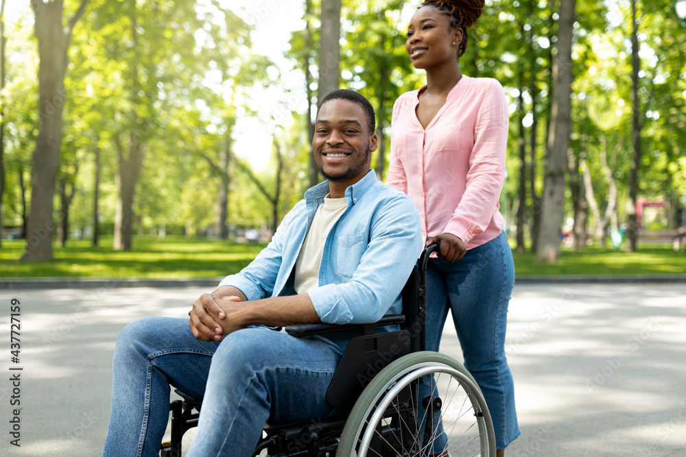 Happy black woman taking her impaired boyfriend in wheelchair for walk at city park - obrazy, fototapety, plakaty 