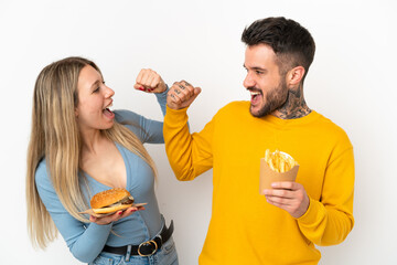 Couple holding hamburger and fried chips over isolated white background celebrating a victory in winner position