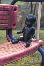 Black Doll Sitting On Dirty Swing At Abandoned Playground