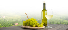 Italian White Wine, Bottle And Glass Of White Wine With Grapes On The Wooden Table, In The Background There Are Vineyards And Hills Of Italian Landscape