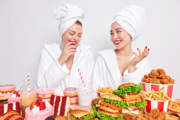 Two happy mixed race women have fun after taking shower stand next to each other wears comfortable bathrobe pose near table full of fast food eats cheat meal do not care about proper nutrition.