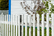 A Narrow White And Blue Wood Cape Cod Siding Exterior Of A Vintage Building With White Trim Around The Window. There's A Country Style White Wooden Picket Fence In The Foreground With Lats And Rails.