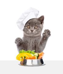 Poster - Kitten wearing chef's hat eats vegetables from a bowl. isolated on white background