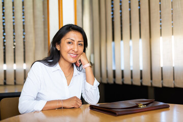 Portrait of confident positive hispanic business woman sitting with briefcase of documents at office table in meeting room.