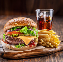 Delicious burger with cola and potato fries on a wooden table with a dark brown background behind. Fast food concept.