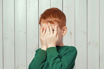 Wall Mural - Close up scared ginger little boy hiding face in palms hands and peering out with one eye between her fingers