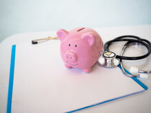 Pink Piggy Bank On White Doctor's Chart And Stethoscope In Copy Space Background. Saving Money For Health And Live Concept