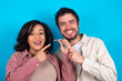 © Roquillo - young couple expecting a baby standing against blue background holding an invisible aligner and pointing at it. Dental healthcare and confidence concept.