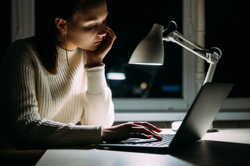 Laptop searching. Boring woman. Internet connection. Distance work. Tired lady leaning hand scrolling notebook in table lamp light dark night room interior.