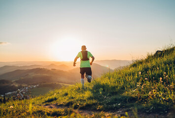 Wall Mural - Active mountain trail runner dressed bright t-shirt with backpack running endurance marathon race by picturesque hills at sunset time back view photo. Sporty active people backlight concept image.