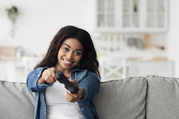 Wall Mural - Playful black woman using joystick to play online video games, sitting on sofa in living room, empty space