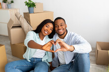 Love Nest. Happy Black Couple Sitting Among Moving Boxes, Making Heart Shape Gesture With Fingers And Smiling To Camera