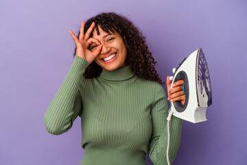 Wall Mural - Young mixed race woman holding an iron isolated on purple background excited keeping ok gesture on eye.