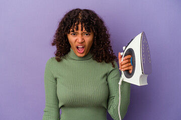 Wall Mural - Young mixed race woman holding an iron isolated on purple background screaming very angry and aggressive.