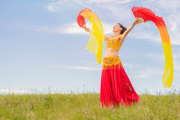 Wall Mural - A young happy woman in national costume with fiery fans in her hands is dancing an incendiary oriental dance on a green lawn against a blue sky. Outdoor activities.